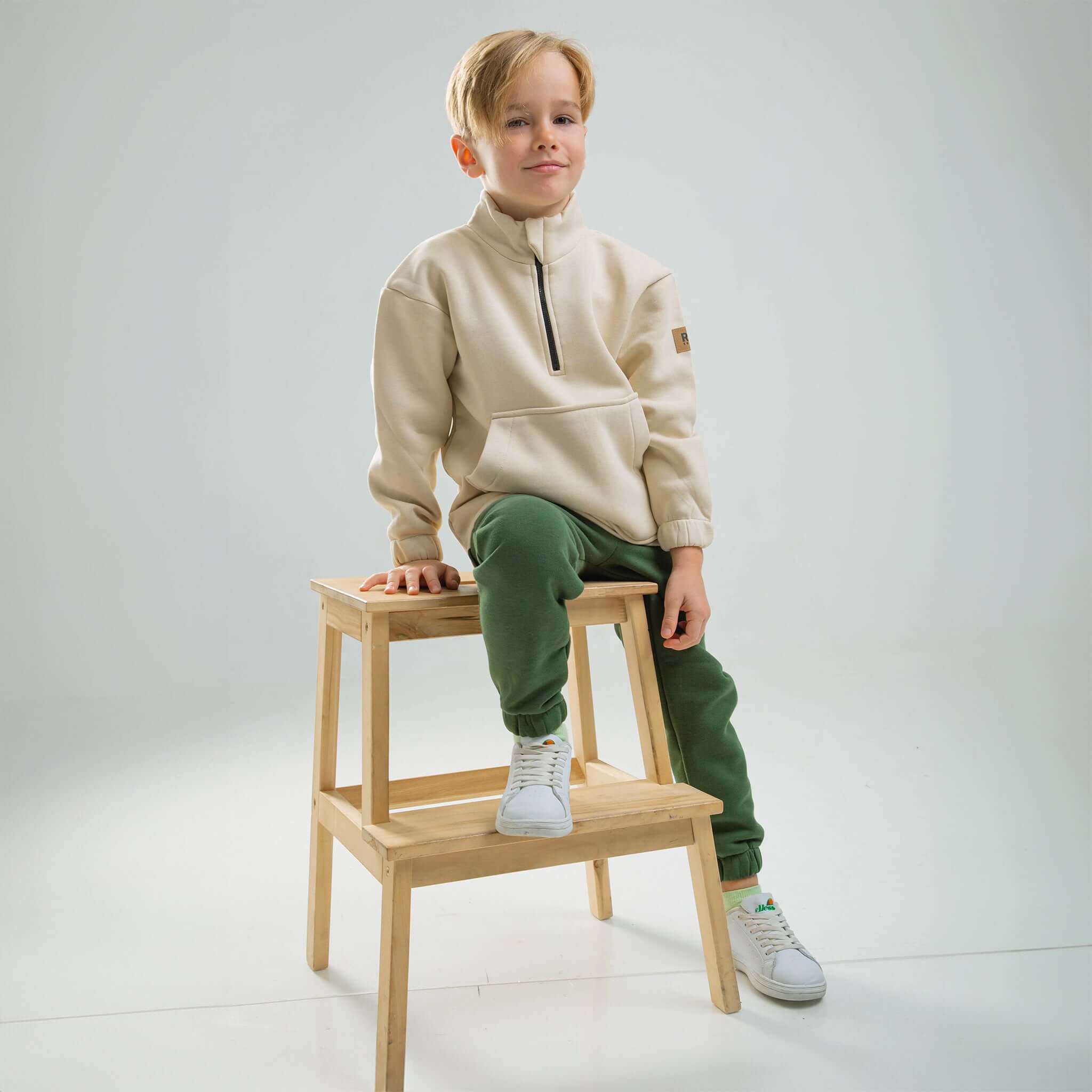Ein Junge sitzt auf einem Hocker und trägt einen RG-Trainingsanzug Ein Junge sitzt auf einem Hocker und trägt einen RG-Trainingsanzug aus Bio-Baumwolle