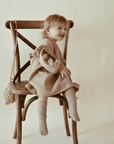 "Ein fröhliches Mädchen sitzt auf dem Stuhl und trägt ein Nehir Kleid aus Bio-Baumwolle."